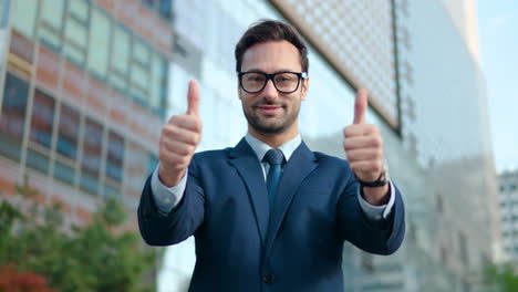 a man in a suit giving two thumbs ups with a big smile on his face standing outside glass skyscrapers in a financial district