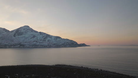 A-slow-drone-shot-flying-over-a-shore-of-the-Kvaloya-Island-in-Norway,-with-snowy-mountain-peaks-in-the-background-and-a-pink-sunset-over-the-horizon