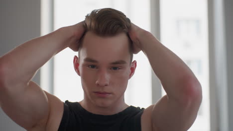 handsome fair skinned young man in black sleeveless top stands before sunlit window mirror, runs fingers through styled hair with focused expression