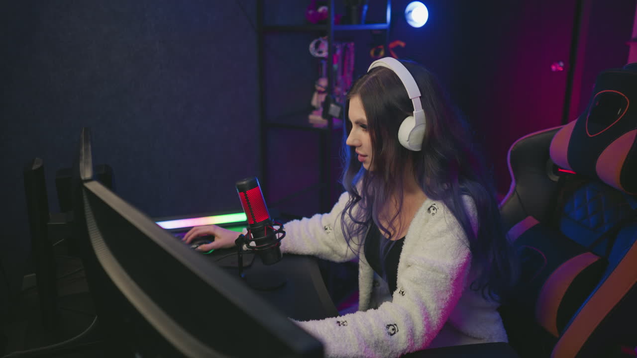 Girl wearing white headphones focused on computer while seated on gaming chair working on creative project in modern setup with microphone, illuminated screen, and decorative shelf in background