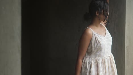 Young-woman-in-a-white-dress-standing-contemplatively-in-a-dimly-lit-concrete-room,-soft-lighting