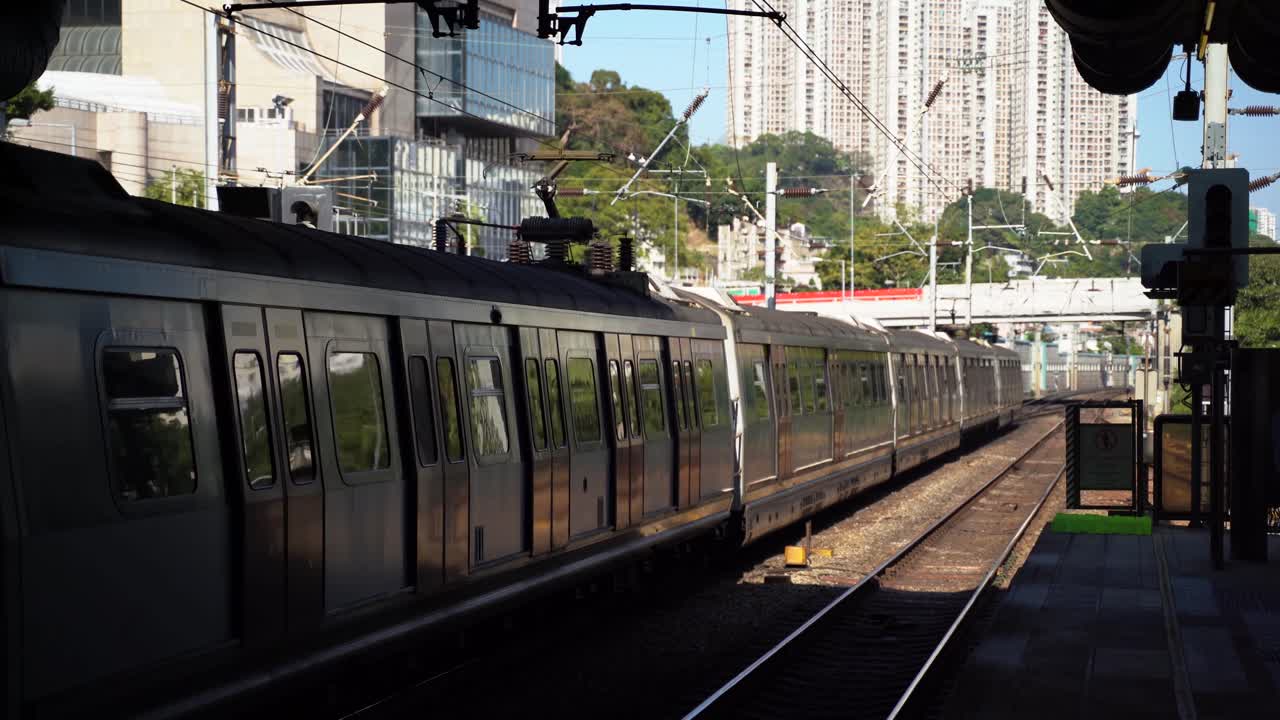 MTR Subway Train in Railway Tracks Departing Station in Hong Kong with Tall Modern Buildings and Hill in Background on a blue sky sunny day