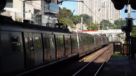 mtr subway train in railway tracks departing station in hong kong with tall modern buildings and hill in background on a blue sky sunny day