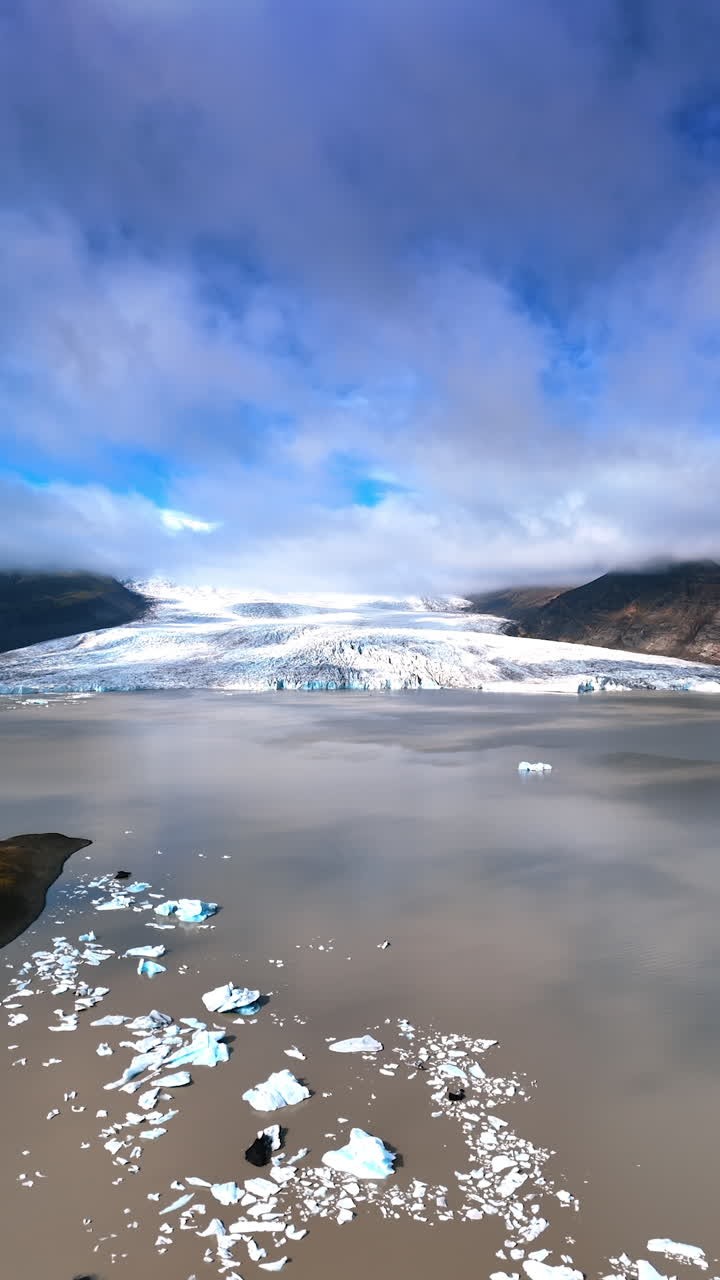 Stunning glacier descends into the waterscape of a cocoa color with some ice pieces floating on the surface. Clouds cover the blue sky. Top view. Vertical video.