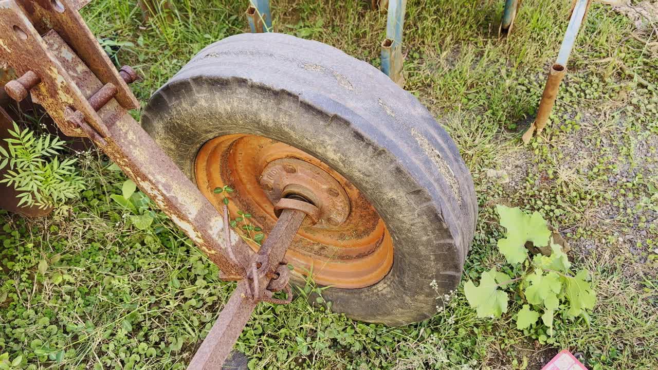 Camera circles around an old rusty wheel of abandoned farm machinery lying on grassy ground, showing texture, decay, and aged metal details