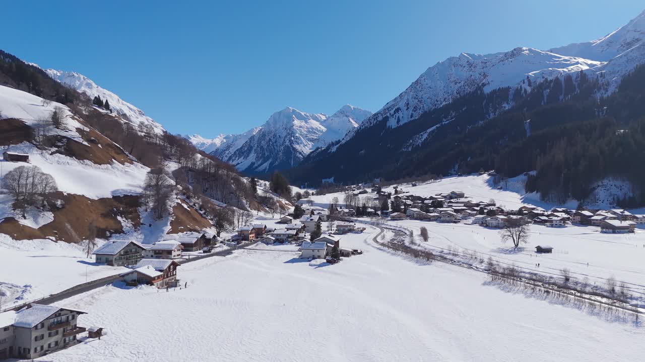 Sorvolo aereo di una valle alpina innevata vicino a Klosters, Svizzera, con case di villaggio, campi aperti e cime montuose spettacolari sotto un sole splendente. Inquadratura ampia. Tempo di vacanza in Svizzera