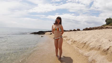 Zoom-in-camera-movement-as-a-brunette-young-woman-in-a-swimsuit-is-smiling-at-the-camera-on-a-sandy-beach-in-Greece-on-a-sunny-day