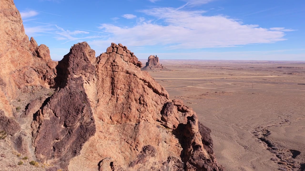 Ripresa drone 4K UHD che scorre lateralmente oltre una parete rocciosa frastagliata, marrone-rossastra in primo piano. Il movimento aereo rivela una vasta, piatta pianura desertica e un imponente rilievo isolato in lontananza.