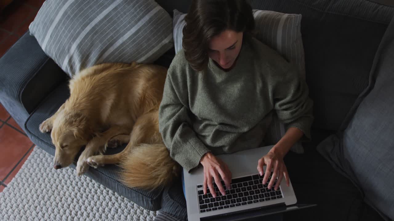 mujer caucásica sonriente usando una computadora portátil trabajando desde casa con su perro de compañía en el sofá a su lado