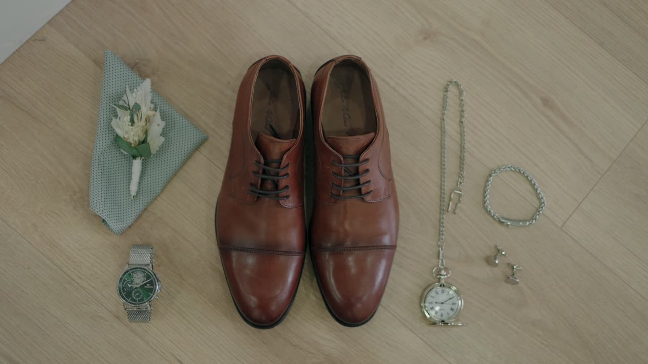 Flat lay of groom's accessories including brown leather shoes, watch, cufflinks, and boutonniere