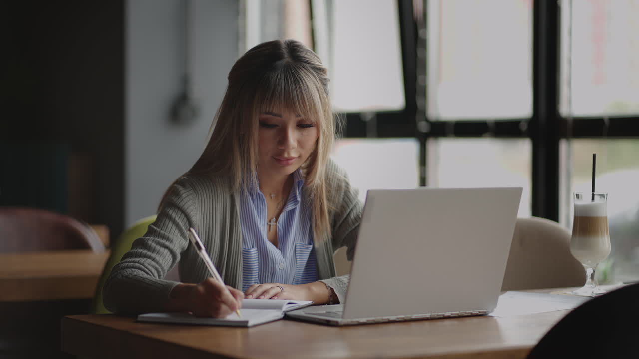 una joven asiática escribe con un bolígrafo mirando la pantalla del portátil sentada en la mesa