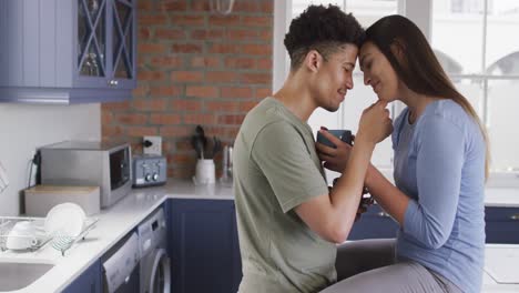 happy biracial couple sitting on countertop in kitchen, drinking coffee