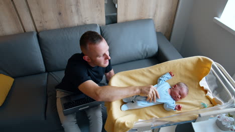 father comforting his newborn son stroking him by the belly. man calms down the child and goes back to work on laptop.