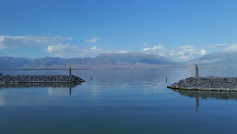 Toma-Aérea-De-La-Entrada-Al-Puerto-Deportivo-De-Saratoga-Springs-En-El-Lago-Utah-Desde-El-Final-De-Un-Pontón-Rojo-Siguiendo-Las-Boyas-Del-Canal-Con-Montañas-Distantes-En-El-Fondo-Reflejándose-En-Las-Aguas-Tranquilas