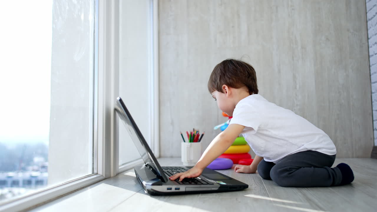 Baby boy pressing keys on the laptop. Kid moves away and sits by the toy pyramid.