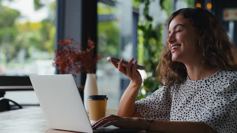 Joven-Empresaria-Sentada-Con-Una-Computadora-Portátil-En-Una-Cafetería-Tomando-Una-Llamada-Por-Teléfono-Móvil-Usando-Un-Micrófono