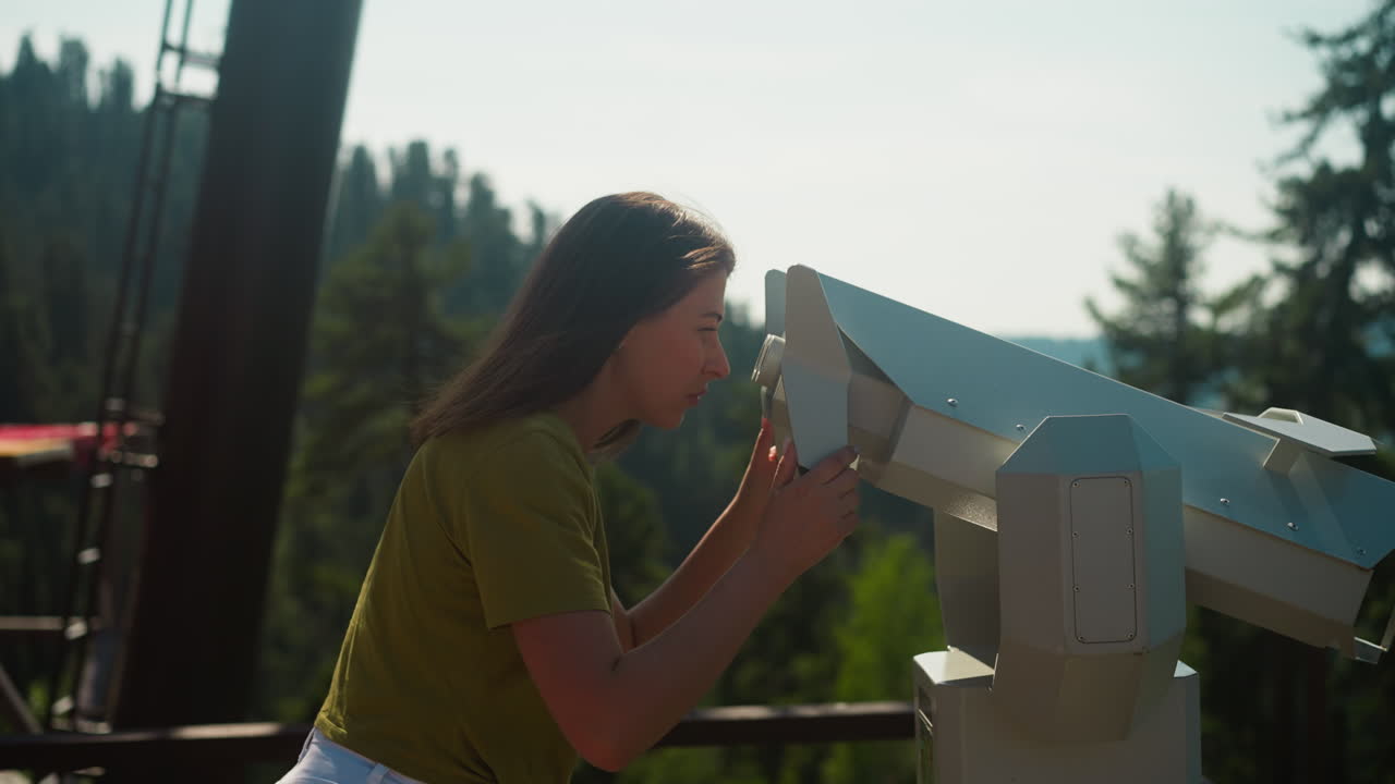 호기심 많은 여자가 관심으로 풍경을 즐기고 관측 갑판에 서있는 안경을 통해보고 있습니다. 두꺼운 나무 숲으로 둘러싸인 관광 지역