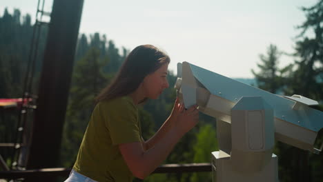 curious woman relishes view with interest and looks through binoculars standing on observation deck. tourist area surrounded by thick coniferous woodland