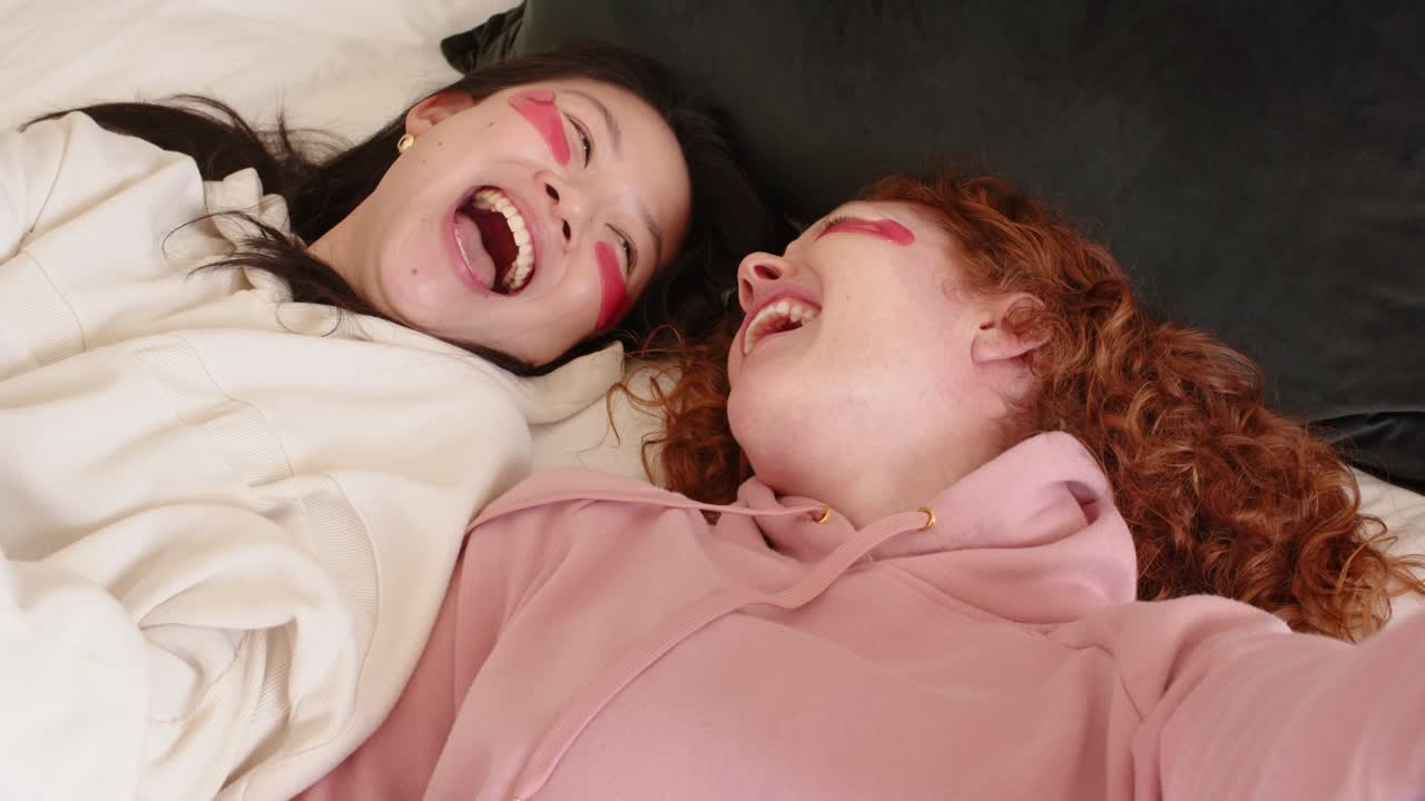 Diverse female friends laughing on bed, left covering mouth, pink eye patches, right posing selfie