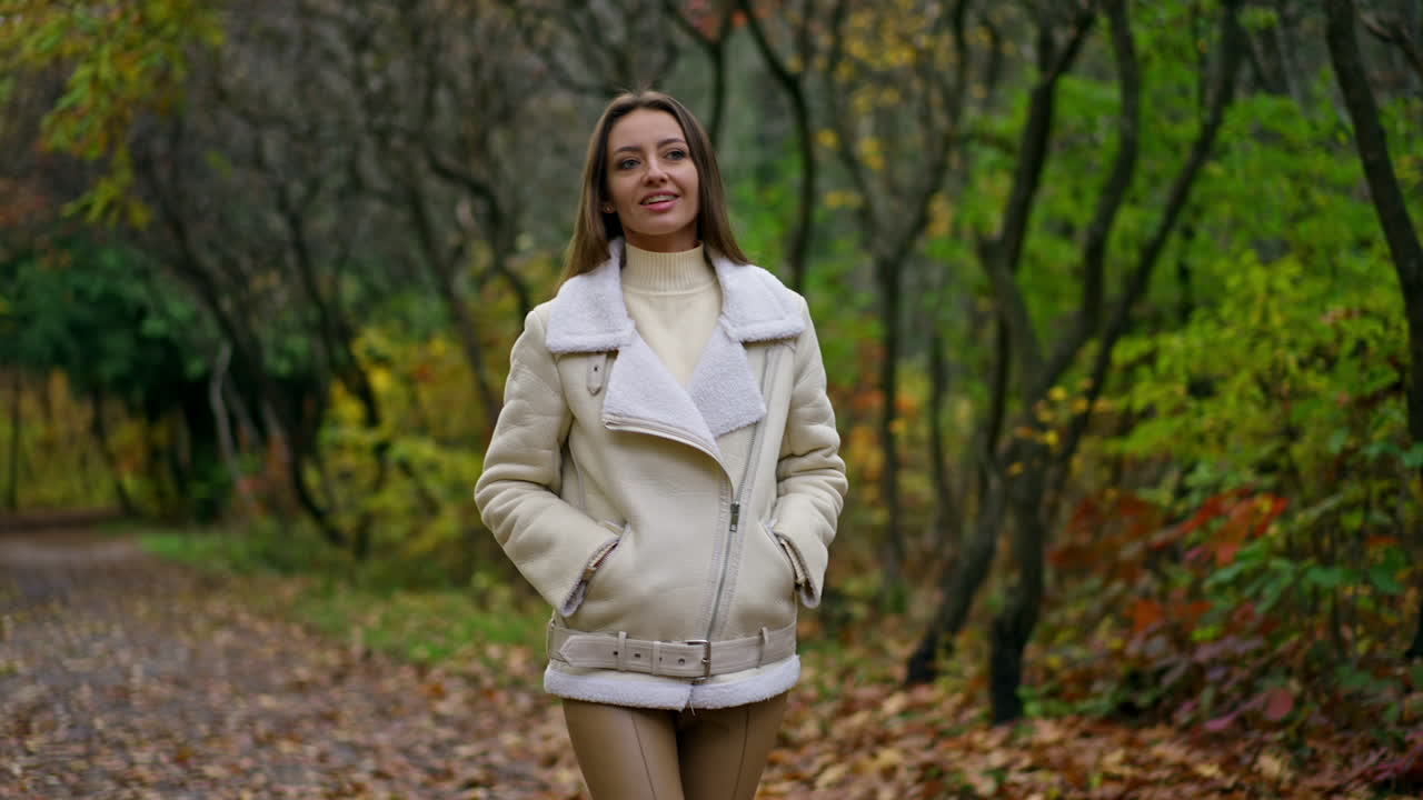 Happy Caucasian lady with long dark hair posing in front of camera in the park. Woman turns smiling to the camera. Autumn nature backdrop.