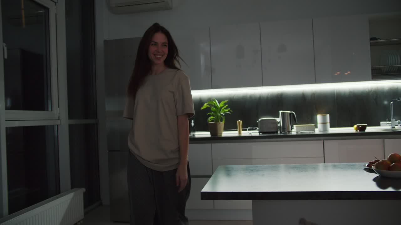 una feliz chica morena en una camiseta beige se acerca a su marido por detrás mientras él en una chaqueta negra y pantalones cortos beige trabaja sentado en el sofá detrás de una computadora portátil gris por la noche en un apartamento de estudio moderno