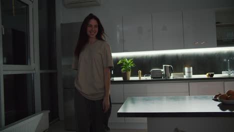 una feliz chica morena en una camiseta beige se acerca a su marido por detrás mientras él en una chaqueta negra y pantalones cortos beige trabaja sentado en el sofá detrás de una computadora portátil gris por la noche en un apartamento de estudio moderno