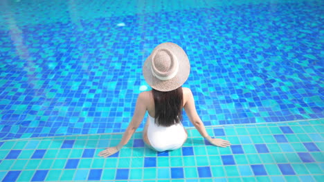 back of exotic sexy woman in white swimsuit and floppy hat sitting on a swimming pool border, fashion concept, full frame