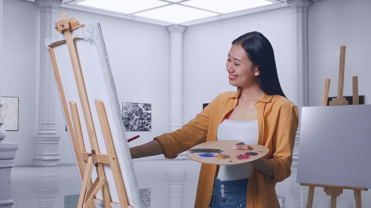 아시아 여성 예술가 가 갤러리에서 오일 페인트와 브러쉬로 캔버스에 그림을 그리는 동안 미소 짓고 카메라에 팔을 고 있습니다.