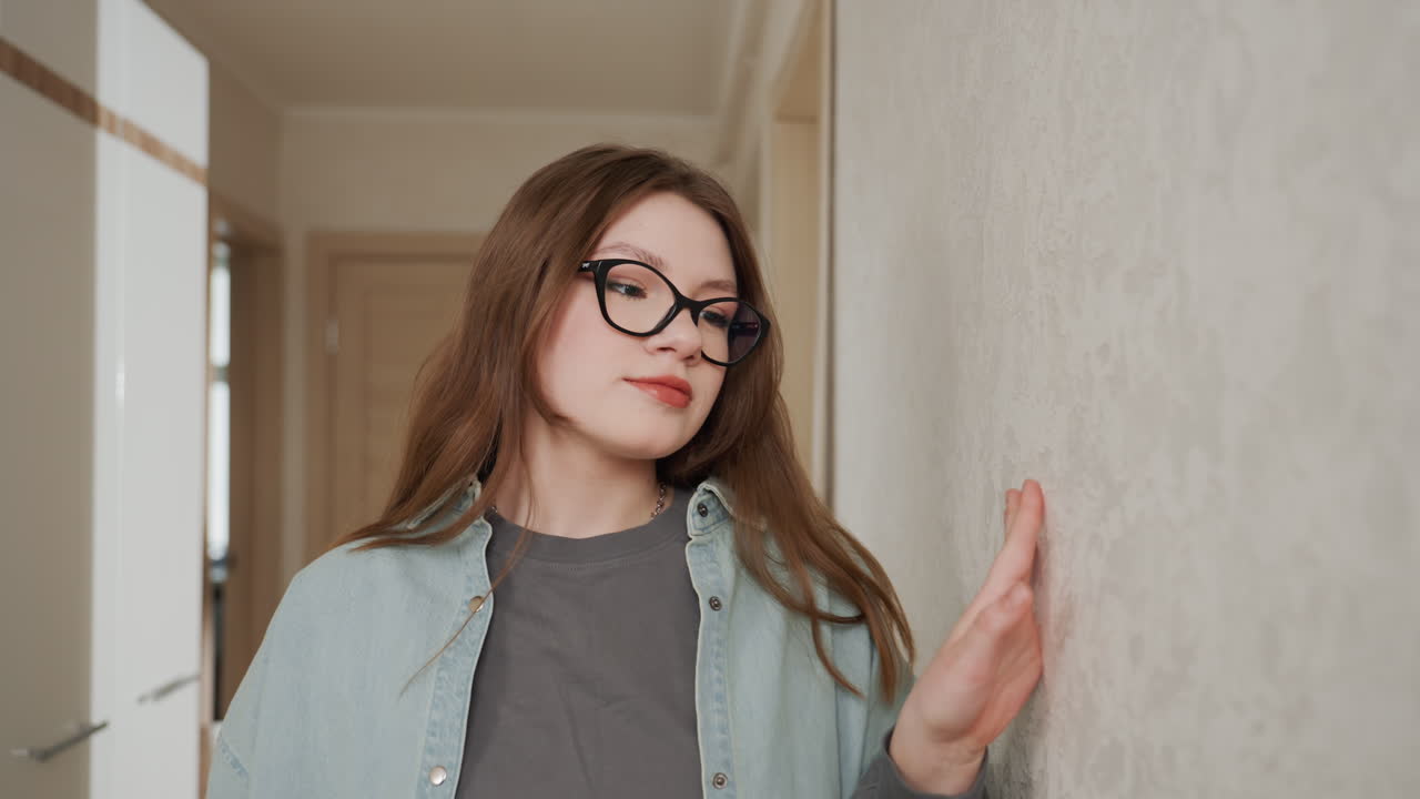 Giovane donna caucasica appoggiata nel corridoio, occhiali, che tocca una parete texturizzata, espressione contemplativa, luce naturale soffusa, abbigliamento casual a strati, atmosfera domestica tranquilla, studentessa che immagina una ristrutturazione