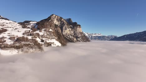 Breathtaking aerial view of thick cloud layer over Swiss alps, covering Amden, Arvenbüel, Weesen, Glarus and Walensee, snowy mountain peaks rise above mist as drone glides forward in winter landscape.