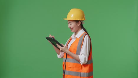 side view of asian female engineer with safety helmet taking note on the tablet while standing in the green screen background studio