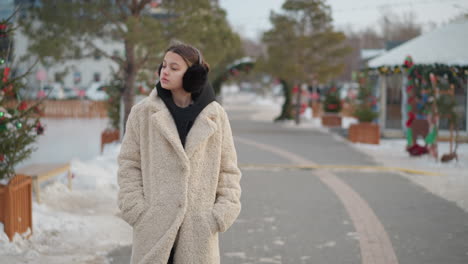 girl wearing warm beige winter coat with black hoodie and earmuffs walks on snowy path, hands tucked in pockets. she gazes at decorated flowers along festive street lined with trees