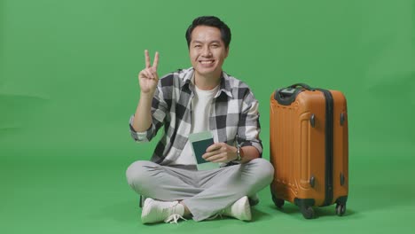 full body of asian male traveler with luggage and passport smiling and showing peace gesture while sitting in the green screen background studio