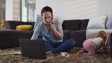 Madre-Caucásica-Usando-Una-Computadora-Portátil-Y-Hablando-Por-Teléfono-Inteligente-Mientras-El-Bebé-Gatea-Por-El-Suelo-En-Casa