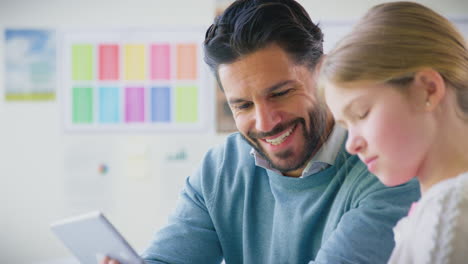 Male-Teacher-With-Female-Student-In-School-Classroom-Sitting-At-Desk-Writing-In-Book-Together