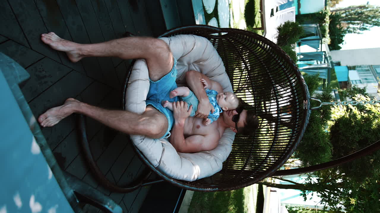 Topless muscular man sits in an outdoor swing chair. Man holding his infant son in hands and kisses him on the head. Vertical video.