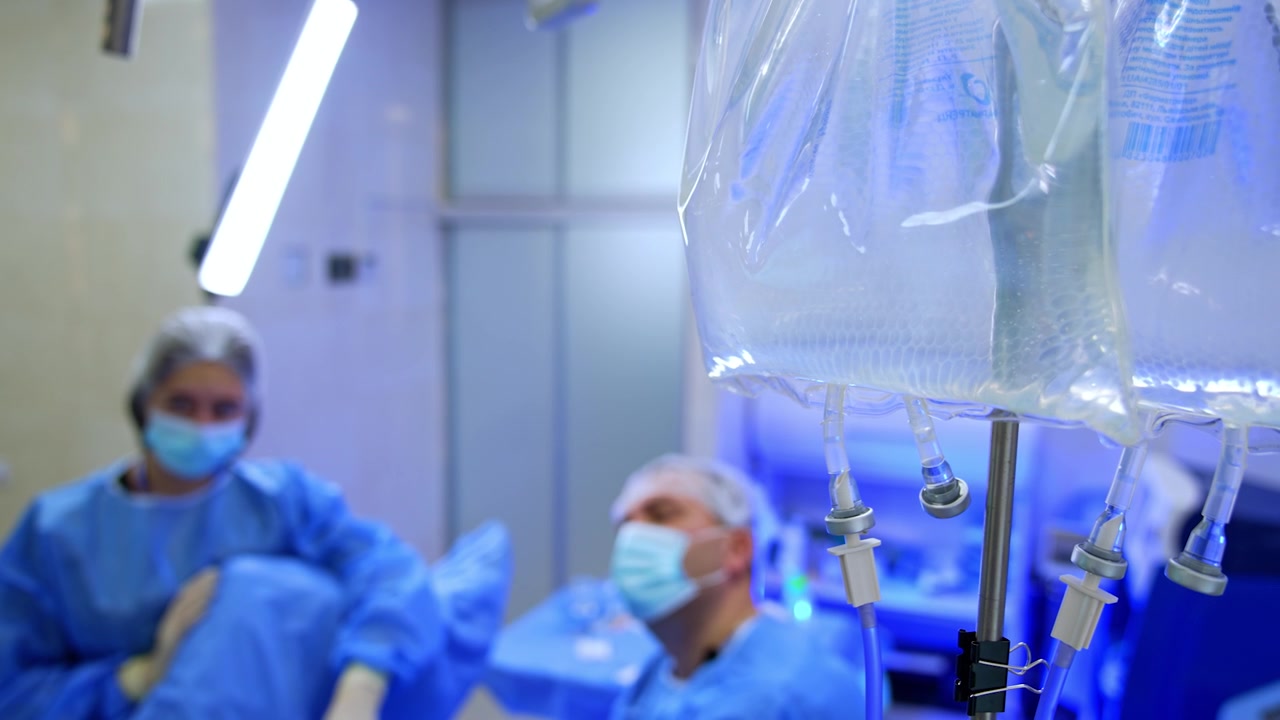 Due sacche di plastica con soluzione fisiologica pendono dal supporto per flebo. Primo piano. Un medico uomo e un'infermiera assistente lavorano su uno sfondo sfocato.