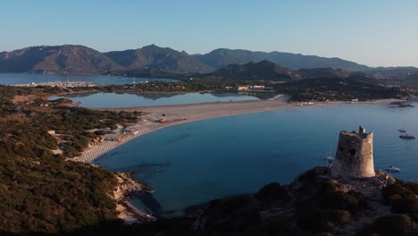 Birds-eye-view-of-Torre-di-Porto-Giunco-tower-standing-tall-on-island-surrounded-by-sea-with-yachts-sailing-with-people-standing-at-coast-in-Italy