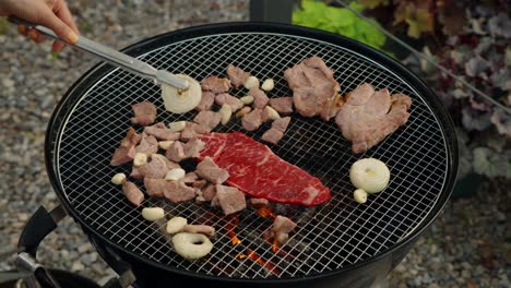 a person uses tongs to cook a fresh, raw, marbled beef steak on hot charcoal grill alongside pork, garlic, and onions during a korean-style garden barbecue