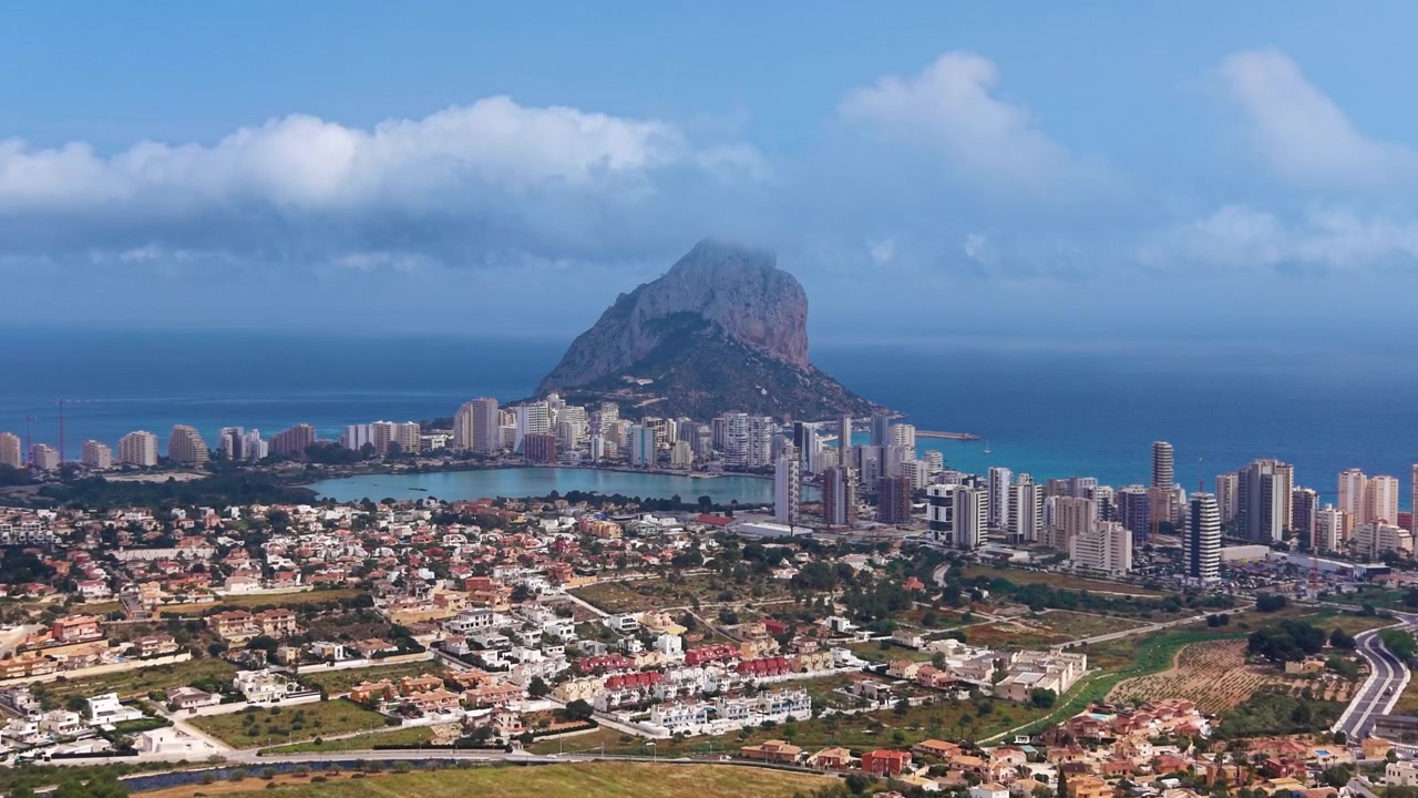 Calpe, Spagna, in una giornata parzialmente nuvolosa che mostra l'iconica formazione rocciosa del Peñón de Ifach che si erge sopra la città costiera, il mare Mediterraneo turchese e lo skyline urbano