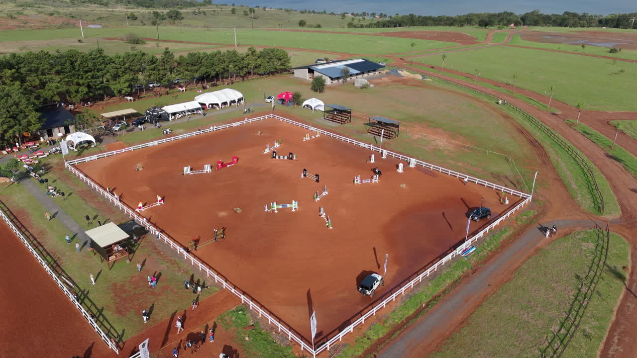 Overhead aerial orbit of horse arena with red sand ring and stables around its perimeter, competitors exhibition practice