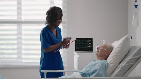 una doctora de piel oscura está hablando con un anciano paciente de sexo masculino acostado en una cama de hospital discutiendo la rehabilitación después de la recuperación.