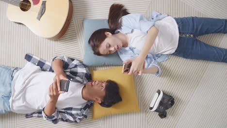 top view of asian teen couple playing smartphone while lying on carpet on the floor at home. phone addicted couple speaking, laughing, and enjoying time together