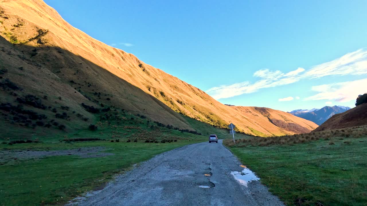 Vehicle travels rural gravel road through sunlit valley, passing trees, bridge, and mountain landscape