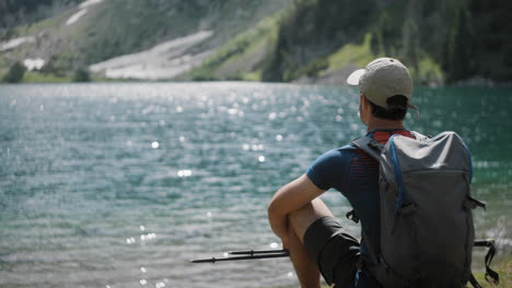 hiker sitting by the mountain lake with the hiking poles and a backpack, mountains in the back, water is glistening