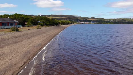 Huillinco-lake-shoreline-with-rustic-homes-and-lush-hills-in-chiloe,-sunny-day,-aerial-view