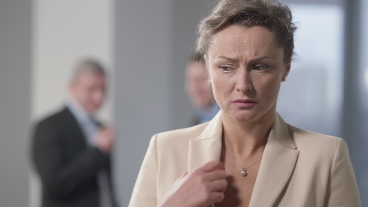 estresada mujer morena con ojos de avellana de pie en el interior pensando con hombres borrosos hablando en el fondo. retrato de una empleada caucásica acosada en la oficina. discriminación y acoso.