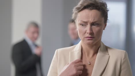 stressed brunette woman with hazel eyes standing indoors thinking with blurred men talking at background. portrait of bullied caucasian female employee in office. discrimination and harassment.