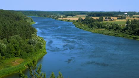 handheld shot of wide river surrounded by green land with tree branches being blownd by light breeze