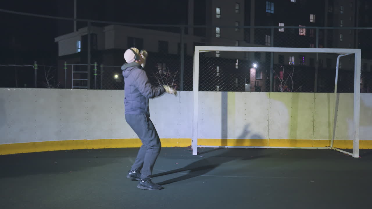 Athlete practicing dynamic football skills, balancing and kicking ball under focused spotlight on outdoor court, urban setting with yellow boundary markings and fencing in background